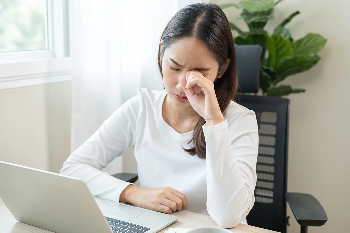 A young woman working on a computer rubs her dry, tired eyes.