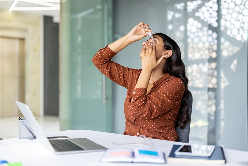 a young woman putting in eye drops for dry eye at work